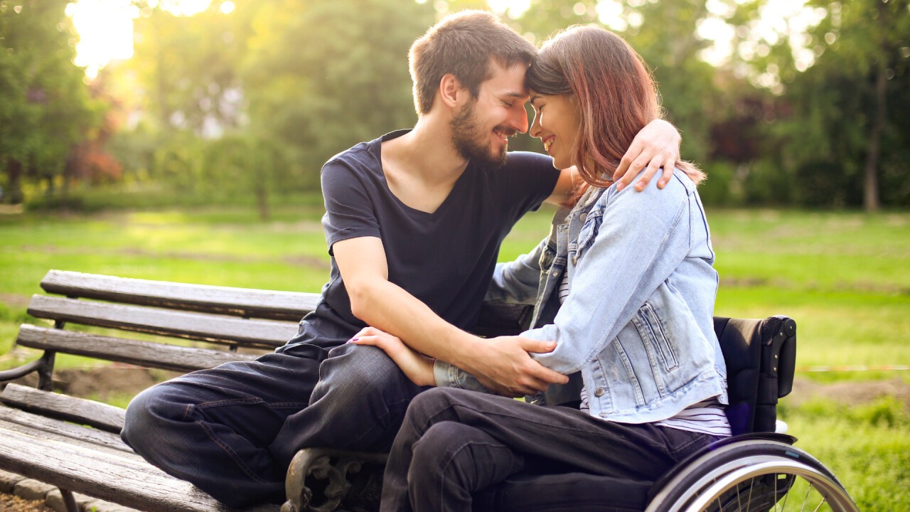 Zwei Menschen, ein Mann links und eine Frau Rechts geniessen die gemeinsame Zeit im Park. Der Mann sizt auf einer Parkbank, die Frau in einem Rollstuhl neben der Bank. Der Mann lehnt sich zur Frau, umarmt sie und beide halten ihre Stirn zusammen, Nase an Nase, so als würden sie sich im nächsten Moment küssen. Sich lachen gemeinsam. Im Hintergrund ist eine Wiese und weiter im Hintergrund sind einige Bäume zu sehen, die zu einem Wald werden.